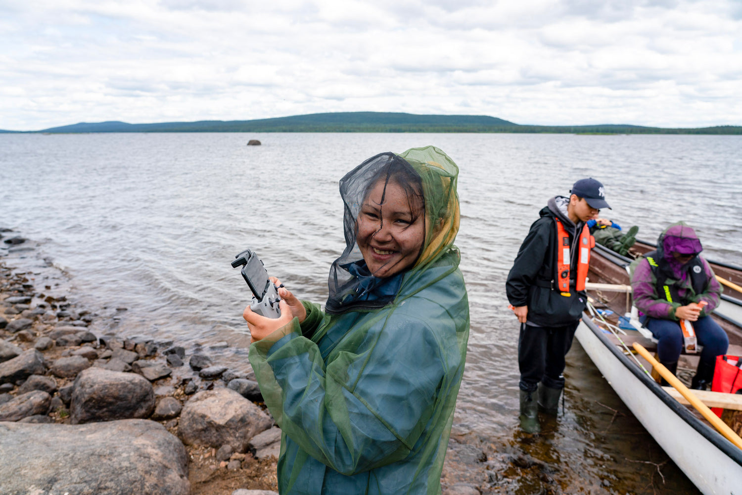 Image of young women holding water testing equipment wearing a bug net at the shore of a lake. Two people behind her. One standing beside a boat the other sitting in the boat.