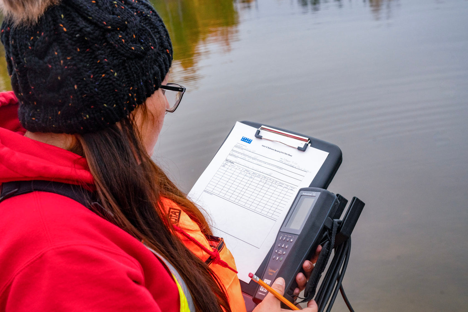 Image of young woman holding a clip board and water testing device standing at the waters edge.