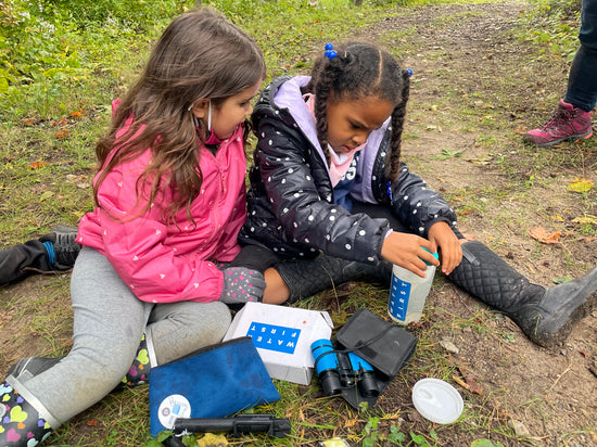 Image of two elementary school girls sitting on the ground outdoors working with a Water First school program kit.